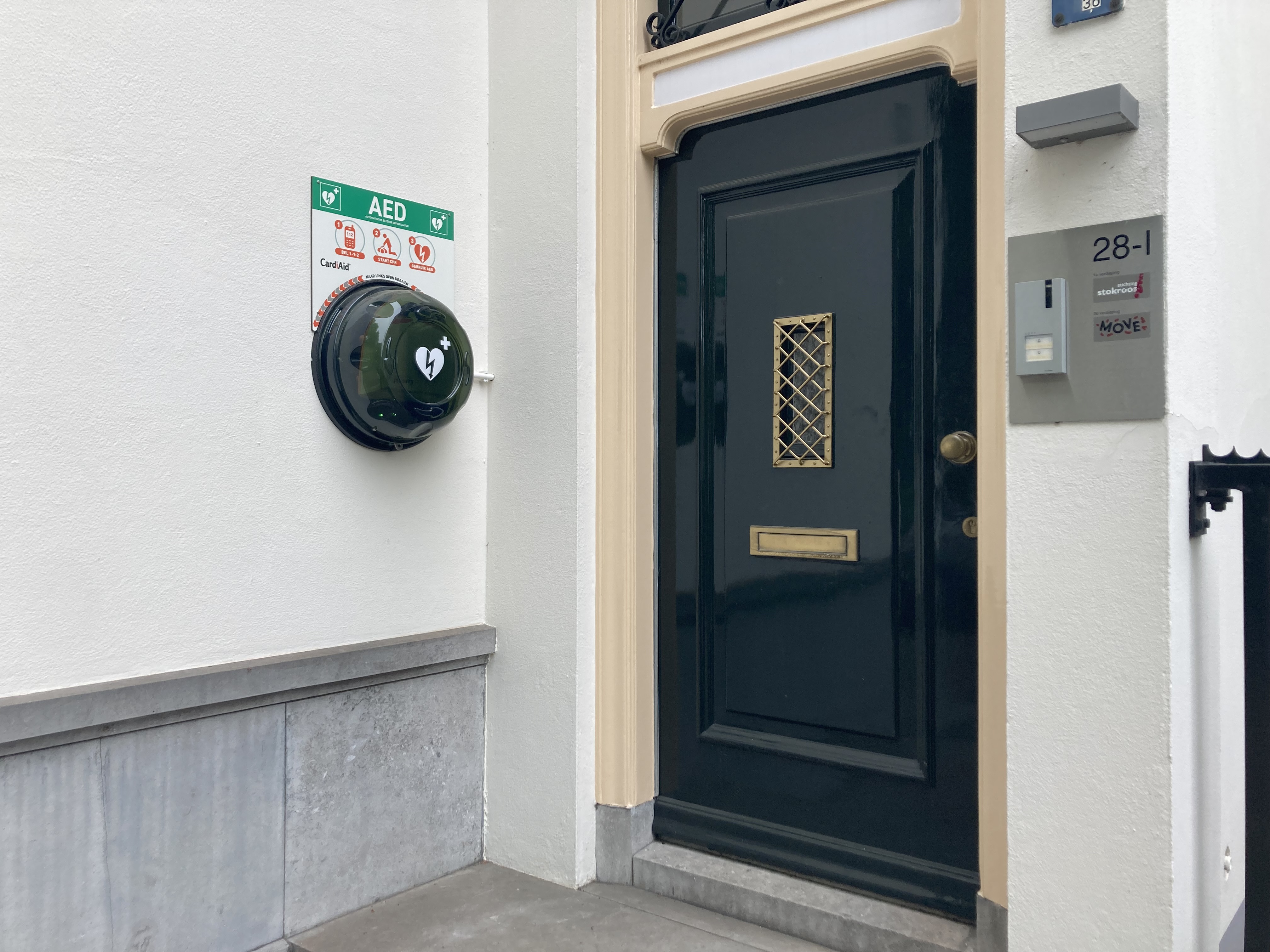 De AED hangt links aan de buitenmuur naast de ingang van het kantoorpand bij nummer 28-1. Op de kast van de AED staat een hart met een bliksemschicht en een plusje. Daarboven hangt een bord met drie pictogrammen hoe de AED gebruikt moet worden